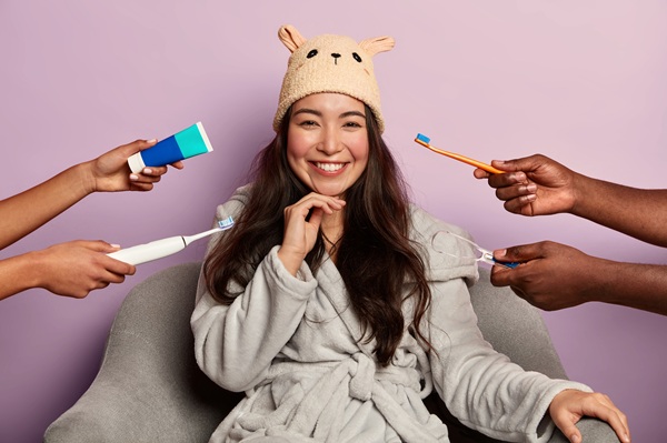Dental care and oral hygiene concept. Positive dark haired young Asian woman wears bathrobe, poses at home, surrounded with dental products, brushes teeth in morning, isolated over violet wall