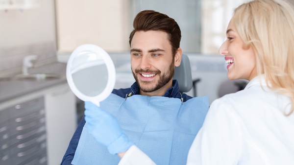 Cheerful doctor dentist holding mirror for happy young man patient sitting in dental chair, doctor and patient checking results of dental treatment, looking at mirror and smiling, panorama, copy space