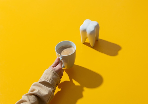 female hand holds cup of coffee next to tooth on yellow background