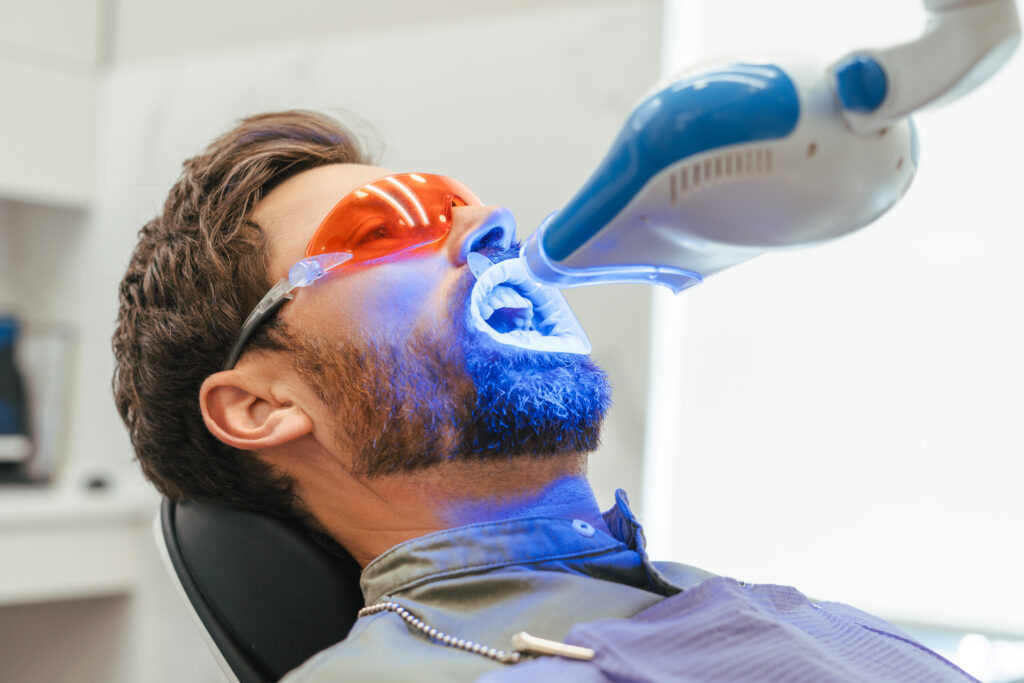 Man receiving Zoom teeth whitening in Sydney, wearing orange protective glasses and a cheek retractor while a blue LED whitening lamp shines on his teeth